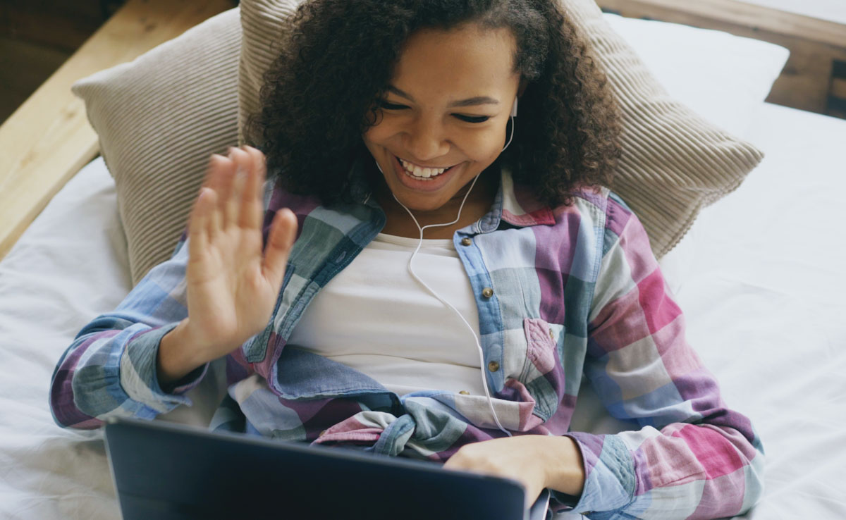 A young woman wearing earphones smiles and waves at her laptop while lying on a bed.