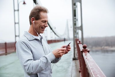 A man wearing earphones smiles at his phone while standing on a bridge.